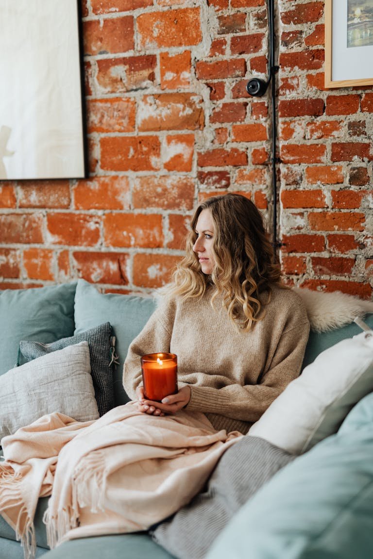 Woman in sweater holding a candle on a sofa against a brick wall, cozy fall atmosphere.